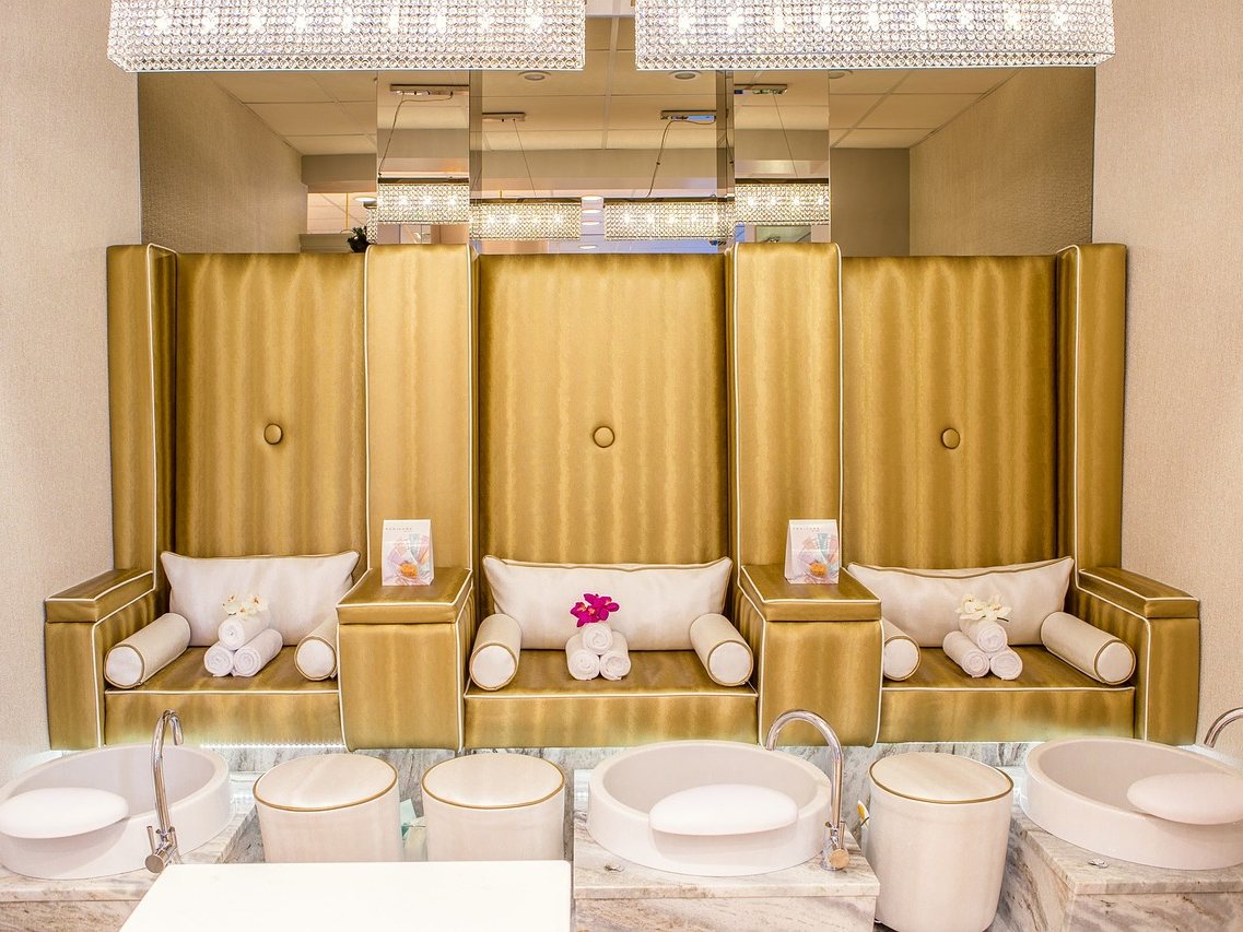 Stock photo of a row of cream and gold upholstered pedicure chairs with rolled towels on the seats and white foot basins below